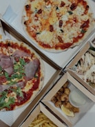 An assortment of food items displayed in open cardboard boxes. Two pizzas feature various toppings including arugula, prosciutto, and cherry tomatoes. Additional boxes contain fried chicken pieces with a lemon wedge and sauce, as well as fries.