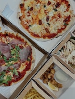 An assortment of food items displayed in open cardboard boxes. Two pizzas feature various toppings including arugula, prosciutto, and cherry tomatoes. Additional boxes contain fried chicken pieces with a lemon wedge and sauce, as well as fries.