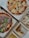 An assortment of food items displayed in open cardboard boxes. Two pizzas feature various toppings including arugula, prosciutto, and cherry tomatoes. Additional boxes contain fried chicken pieces with a lemon wedge and sauce, as well as fries.