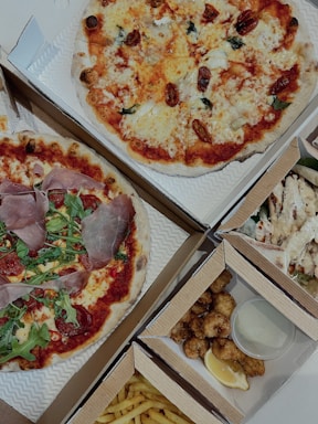 An assortment of food items displayed in open cardboard boxes. Two pizzas feature various toppings including arugula, prosciutto, and cherry tomatoes. Additional boxes contain fried chicken pieces with a lemon wedge and sauce, as well as fries.