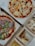 An assortment of food items displayed in open cardboard boxes. Two pizzas feature various toppings including arugula, prosciutto, and cherry tomatoes. Additional boxes contain fried chicken pieces with a lemon wedge and sauce, as well as fries.