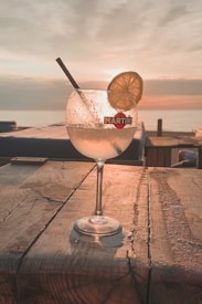 A cocktail glass filled with an iced beverage rests on a wooden surface, garnished with a slice of lemon and a straw. The background features a serene, colorful sunset over a calm sea, creating a warm and tranquil atmosphere.
