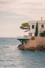 A Mediterranean-style villa is perched on rocky cliffs by the sea. The building is white with arched windows and surrounded by lush greenery, including a prominent tree. A sailboat is visible in the distance on the ocean under a cloudy sky.