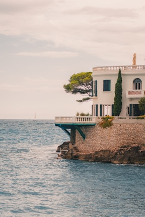 A Mediterranean-style villa is perched on rocky cliffs by the sea. The building is white with arched windows and surrounded by lush greenery, including a prominent tree. A sailboat is visible in the distance on the ocean under a cloudy sky.