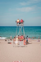Two lifeguards sit on an elevated chair, overlooking a busy beach with several people enjoying the water and sand. Various beach items like towels and umbrellas can be seen, along with a few individuals near the shoreline.