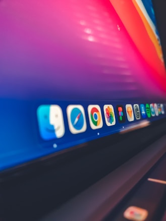 A close-up view of a computer screen displaying a dock with various application icons including Finder, Safari, Chrome, Photos, Spotify, and others. The background is a gradient of pink, orange, and blue colors.