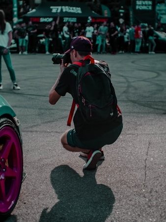 A photographer crouches on an asphalt surface, focusing through a camera held up to their eye. They are wearing a backpack with red straps and a cap, dressed in casual attire with sneakers. The foreground features a purple car wheel, while in the background, a crowd of people appear to be watching something out of frame. Several large advertising banners and tents are also visible, suggesting a public event.