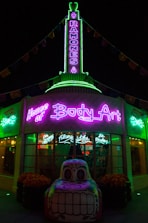 A building illuminated with vibrant neon lights, featuring signage for 'House of Body Art' and 'Ramones' in bright green and purple. In front of the building, there is a decorative car sculpture with a colorful, stylized face. The shop windows display signs for services like 'Flame Jobs' and 'Air Brushing'.