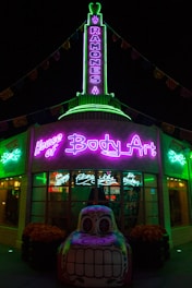 A building illuminated with vibrant neon lights, featuring signage for 'House of Body Art' and 'Ramones' in bright green and purple. In front of the building, there is a decorative car sculpture with a colorful, stylized face. The shop windows display signs for services like 'Flame Jobs' and 'Air Brushing'.