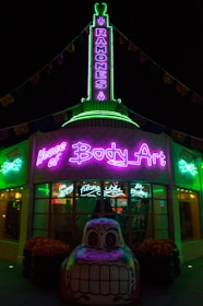 A building illuminated with vibrant neon lights, featuring signage for 'House of Body Art' and 'Ramones' in bright green and purple. In front of the building, there is a decorative car sculpture with a colorful, stylized face. The shop windows display signs for services like 'Flame Jobs' and 'Air Brushing'.