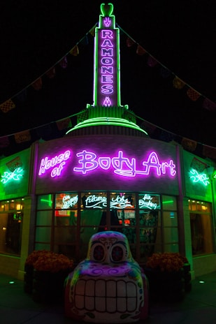 A building illuminated with vibrant neon lights, featuring signage for 'House of Body Art' and 'Ramones' in bright green and purple. In front of the building, there is a decorative car sculpture with a colorful, stylized face. The shop windows display signs for services like 'Flame Jobs' and 'Air Brushing'.