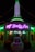 A building illuminated with vibrant neon lights, featuring signage for 'House of Body Art' and 'Ramones' in bright green and purple. In front of the building, there is a decorative car sculpture with a colorful, stylized face. The shop windows display signs for services like 'Flame Jobs' and 'Air Brushing'.