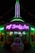 A building illuminated with vibrant neon lights, featuring signage for 'House of Body Art' and 'Ramones' in bright green and purple. In front of the building, there is a decorative car sculpture with a colorful, stylized face. The shop windows display signs for services like 'Flame Jobs' and 'Air Brushing'.