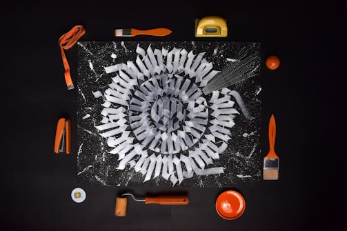 A mandala-like geometric pattern is featured on a dark background, surrounded by various orange-colored tools and objects. Items such as a stapler, tape measure, paintbrushes, a paint roller, and a small bowl complement the design. The central artwork consists of intricate white shapes arranged radially, resembling a spiral.