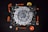 A mandala-like geometric pattern is featured on a dark background, surrounded by various orange-colored tools and objects. Items such as a stapler, tape measure, paintbrushes, a paint roller, and a small bowl complement the design. The central artwork consists of intricate white shapes arranged radially, resembling a spiral.