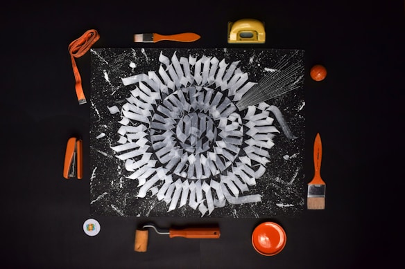 A mandala-like geometric pattern is featured on a dark background, surrounded by various orange-colored tools and objects. Items such as a stapler, tape measure, paintbrushes, a paint roller, and a small bowl complement the design. The central artwork consists of intricate white shapes arranged radially, resembling a spiral.