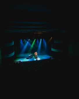 A musician is seated on a dimly lit stage, playing an acoustic guitar while surrounded by colorful stage lights. The lighting is a mix of blue, green, and purple hues, casting a moody atmosphere. The stage is mostly empty, with a piano or keyboard nearby, and the audience area is dark and appears unoccupied.