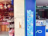 A shop display with an assortment of colorful backpacks and footwear on shelves. The adjacent store features promotional posters for a 20MP Softlight Camera smartphone by Vivo. Inside, there's a clean, modern interior with lit displays.
