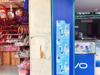 A shop display with an assortment of colorful backpacks and footwear on shelves. The adjacent store features promotional posters for a 20MP Softlight Camera smartphone by Vivo. Inside, there's a clean, modern interior with lit displays.