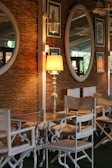 A cozy indoor setting featuring a rustic woven wall with decorative elements. Two large oval mirrors and framed botanical artwork adorn the wall. A warm lamp with a white stand sits on a wooden table surrounded by classic white chairs with canvas seating. The ambiance is enhanced by soft light and subtle reflections.