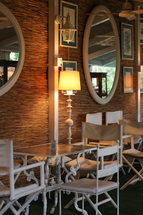 A cozy indoor setting featuring a rustic woven wall with decorative elements. Two large oval mirrors and framed botanical artwork adorn the wall. A warm lamp with a white stand sits on a wooden table surrounded by classic white chairs with canvas seating. The ambiance is enhanced by soft light and subtle reflections.