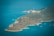 A lush, green island surrounded by the clear blue waters of the sea. Several sailboats are visible in the water, providing a sense of tranquility and leisure. The coastline features a mix of rocky and sandy areas, creating a picturesque landscape.