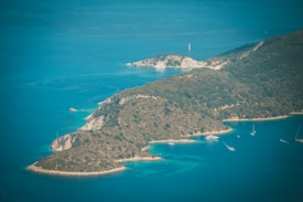 A lush, green island surrounded by the clear blue waters of the sea. Several sailboats are visible in the water, providing a sense of tranquility and leisure. The coastline features a mix of rocky and sandy areas, creating a picturesque landscape.