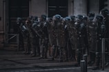 A group of riot police officers stands in a line with shields and helmets, seemingly prepared for crowd control. They are positioned in front of a building in an urban environment under dim lighting.