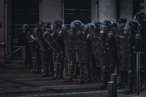 A group of riot police officers stands in a line with shields and helmets, seemingly prepared for crowd control. They are positioned in front of a building in an urban environment under dim lighting.