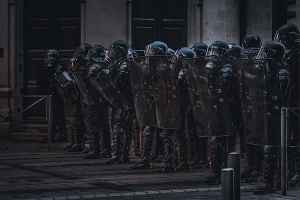 A group of riot police officers stands in a line with shields and helmets, seemingly prepared for crowd control. They are positioned in front of a building in an urban environment under dim lighting.
