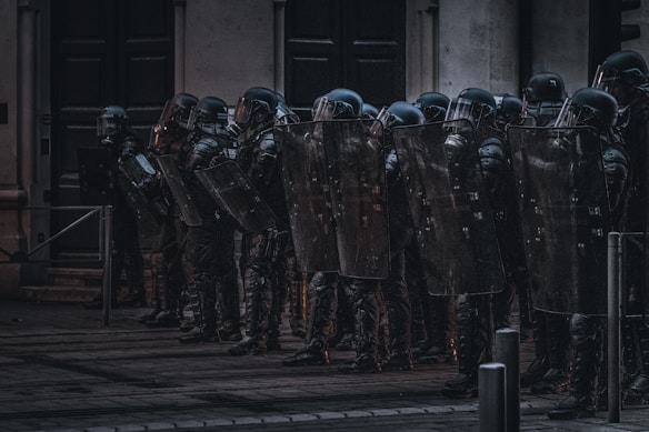 A group of riot police officers stands in a line with shields and helmets, seemingly prepared for crowd control. They are positioned in front of a building in an urban environment under dim lighting.