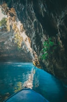 A serene cave with turquoise water and sunlight streaming through an opening at the top, illuminating the rocky walls and reflecting off the water. A small portion of a blue boat is visible in the lower foreground, suggesting a peaceful exploration.