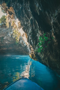 A serene cave with turquoise water and sunlight streaming through an opening at the top, illuminating the rocky walls and reflecting off the water. A small portion of a blue boat is visible in the lower foreground, suggesting a peaceful exploration.