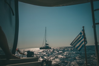 A sailboat glides across a sunlit ocean, casting a shimmering reflection on the water. A Greek flag flutters from a nearby vessel, suggesting a coastal location under a clear sky.