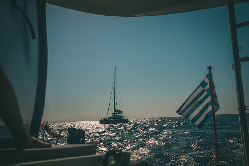 Sailboat gently gliding past sunlit, whitewashed Greek island coastline with clear blue water.