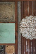 The image displays a textured wall with an assortment of decorative tiles and metal panels. The tiles include ornate floral and abstract carvings, framed by rusted and weathered wooden sections. One tile is embossed with a large floral pattern, while others feature aged paint and patina.