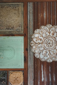 The image displays a textured wall with an assortment of decorative tiles and metal panels. The tiles include ornate floral and abstract carvings, framed by rusted and weathered wooden sections. One tile is embossed with a large floral pattern, while others feature aged paint and patina.
