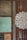 The image displays a textured wall with an assortment of decorative tiles and metal panels. The tiles include ornate floral and abstract carvings, framed by rusted and weathered wooden sections. One tile is embossed with a large floral pattern, while others feature aged paint and patina.