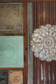 The image displays a textured wall with an assortment of decorative tiles and metal panels. The tiles include ornate floral and abstract carvings, framed by rusted and weathered wooden sections. One tile is embossed with a large floral pattern, while others feature aged paint and patina.