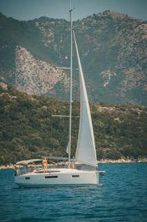 A sailboat with a white hull and large sail is cruising on a body of water. Two people are visible on deck, one standing near the edge and another near the mast. The backdrop features towering, green-covered mountains under a clear sky.
