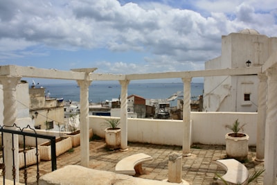 A coastal view with whitewashed buildings and an open terrace with columns overlooking the sea. The sky is partly cloudy, creating a serene atmosphere. Small plants in pots occupy parts of the terrace, adding a touch of greenery.
