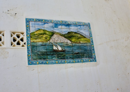 A framed ceramic tile mural is mounted on a textured white wall. The mural depicts a scenic view of a coastal landscape with a sailboat in the foreground and mountainous terrain in the background. The mural is bordered with a decorative floral design.