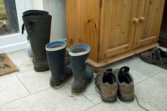 A pair of black rubber boots with blue trim and some grass clinging to them sit on a tiled floor, next to a pair of brown hiking shoes and sandals. A wooden cabinet stands nearby, and a doormat can be seen partially in the background.