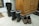 A pair of black rubber boots with blue trim and some grass clinging to them sit on a tiled floor, next to a pair of brown hiking shoes and sandals. A wooden cabinet stands nearby, and a doormat can be seen partially in the background.