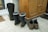 A pair of black rubber boots with blue trim and some grass clinging to them sit on a tiled floor, next to a pair of brown hiking shoes and sandals. A wooden cabinet stands nearby, and a doormat can be seen partially in the background.