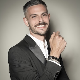 A man with a neatly groomed beard and short haircut is wearing a black suit with a white shirt. He smiles confidently while holding up his right hand, displaying several bracelets on his wrist.
