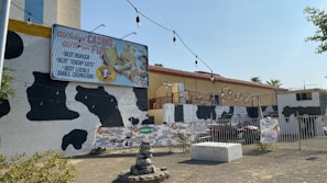 A building exterior with a cow print design on its facade. A large sign advertises 'Old School Casino Old School Fun' and mentions 'Best Burger, Best Cheap Eats, Best Locals, Small Casino/Bar.' The scene includes outdoor string lights, several advertising signs for beverages, and a small water fountain with rocks in the foreground. The building has a flat roof and a fence surrounds the area.