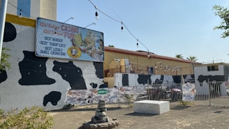 A building exterior with a cow print design on its facade. A large sign advertises 'Old School Casino Old School Fun' and mentions 'Best Burger, Best Cheap Eats, Best Locals, Small Casino/Bar.' The scene includes outdoor string lights, several advertising signs for beverages, and a small water fountain with rocks in the foreground. The building has a flat roof and a fence surrounds the area.