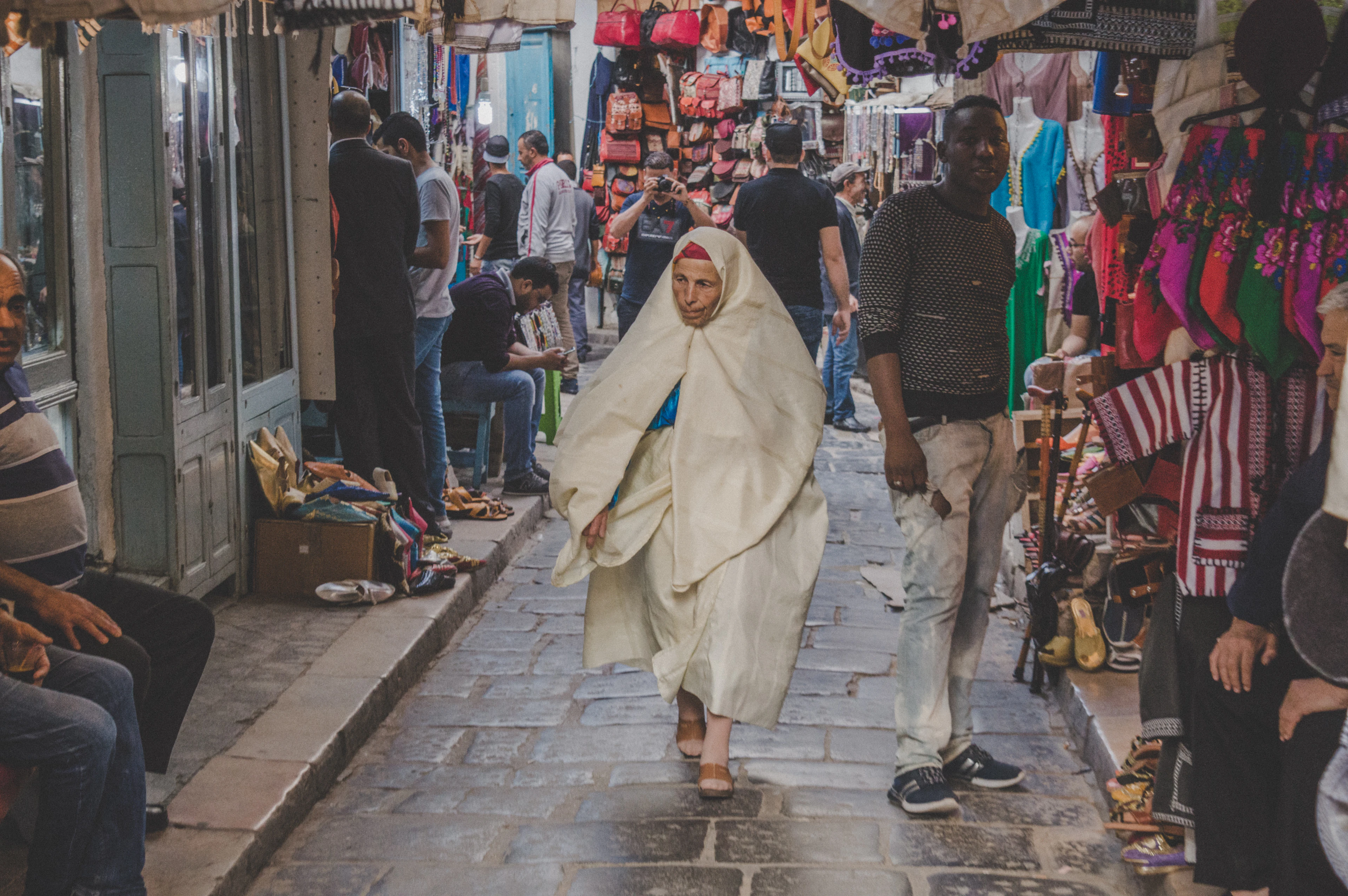 A woman in white robes walks a narrow medina lane lined with market stalls selling scarves and bags.