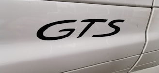 The image features a close-up view of the word 'GTS' in black lettering on the side panel of a white vehicle. The background shows smooth, glossy paint with light reflections and some visible panel lines.